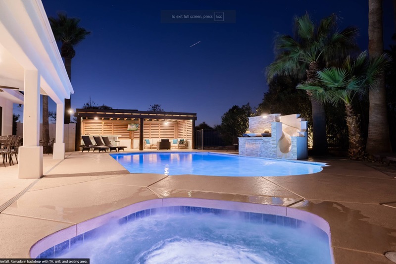 Nighttime view of a Scottsdale vacation home with a lit swimming pool, hot tub, outdoor seating, and palm trees under a clear sky.