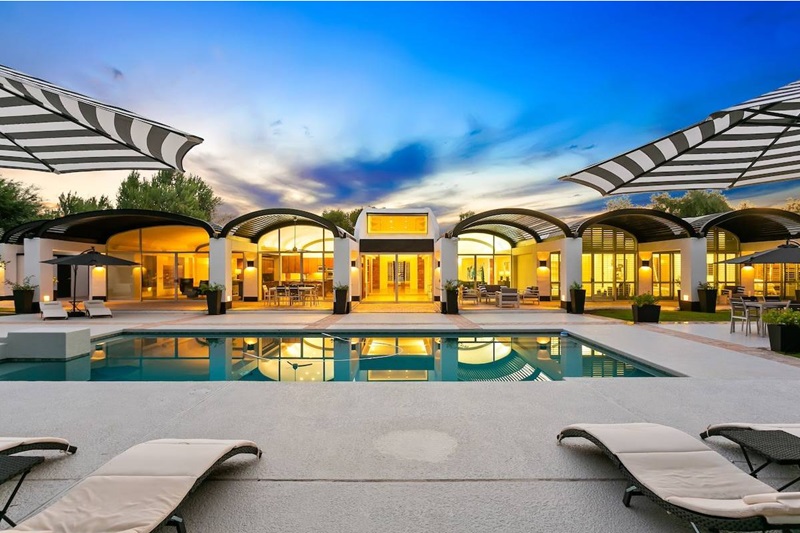 Luxurious Scottsdale vacation home with a modern design, illuminated interior, outdoor pool, and stylish lounge chairs at sunset.