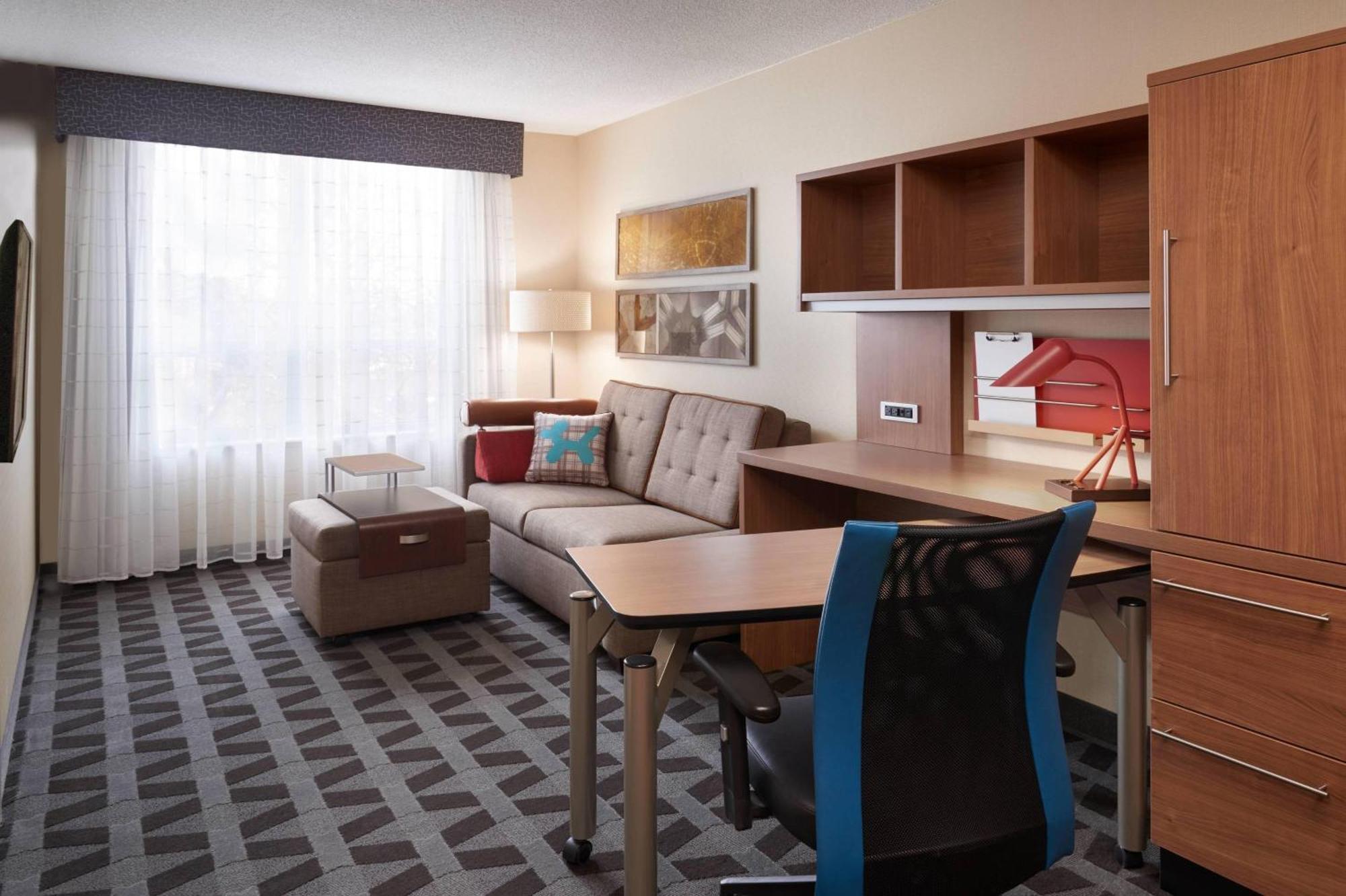 Interior of a hotel room at Townplace Suites Windsor, featuring a sofa, desk, chair, and window with curtains.