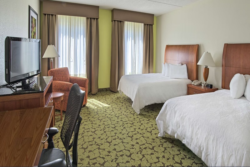 Hotel room with two beds, a TV, a desk, an armchair, and large windows with curtains, in a Hilton Garden Inn Huntsville.