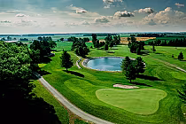 Aerial view of Oxford Hills golf course with lush green fairways, a small pond, trees, and open fields under a partly cloudy sky.