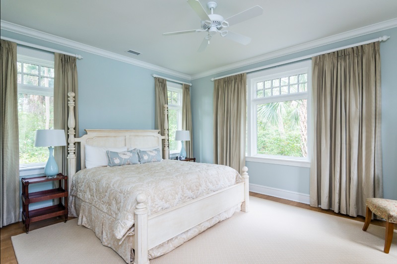 Private Homes - Kiawah Island Resort Bright bedroom with a white bed, beige bedding, large windows with beige curtains, and a view of lush greenery outside.