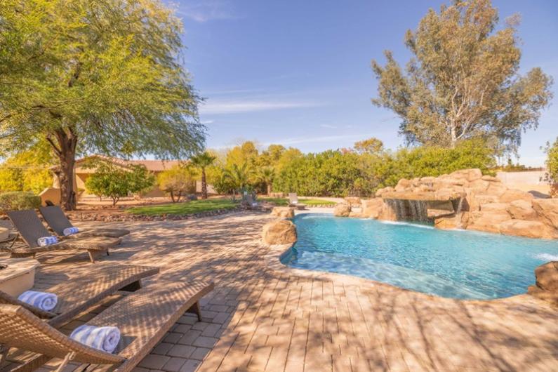 Luxury Scottsdale vacation home with a swimming pool, lounge chairs, lush trees, and a sunny sky, perfect for golfing and relaxation.