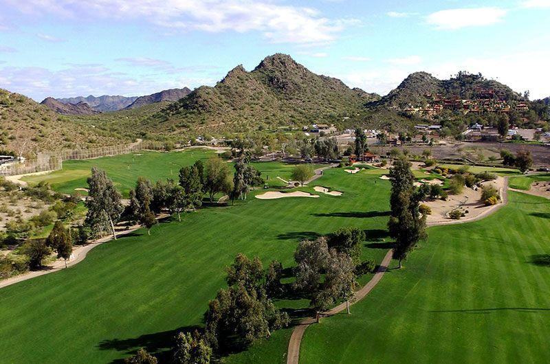 A scenic golf course in Arizona with lush green fairways, trees, sand traps, and mountainous background under a blue sky.