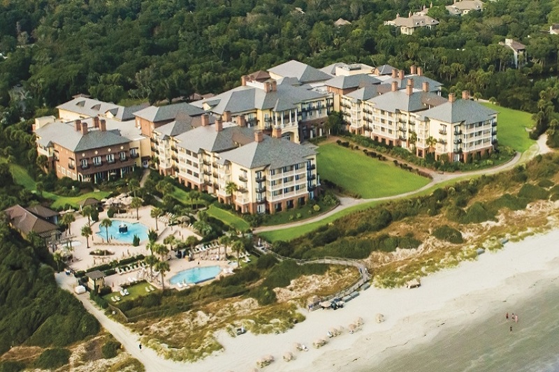 The Sanctuary - Kiawah Island Resort A luxury resort on Kiawah Island with multiple buildings, a swimming pool, lush greenery, and a beach with umbrellas and visitors.