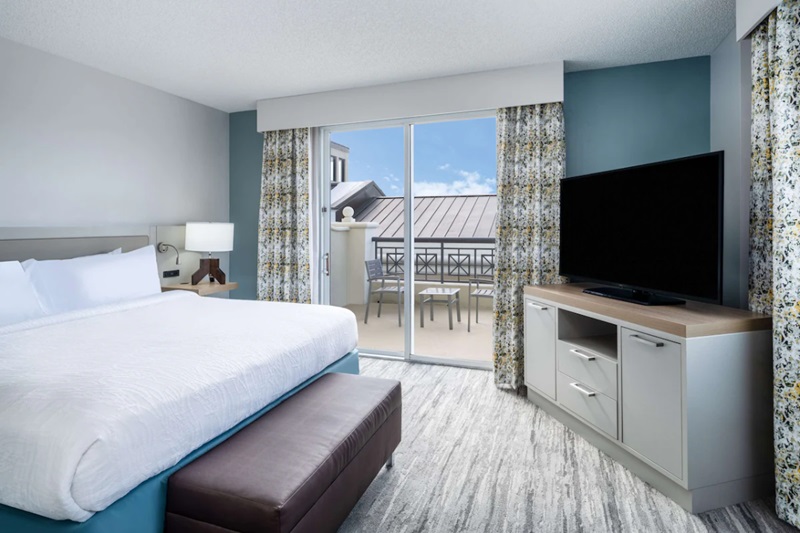 Modern hotel room with a balcony, featuring a large bed, a TV, and a view of the balcony with outdoor seating, in a luxury pet-friendly hotel.