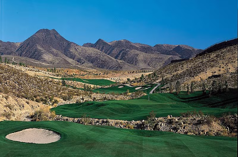 Scenic view of a golf course with green fairways, sand bunkers, and mountainous desert landscape under a clear blue sky.