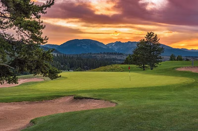 Scenic golf course at sunset in the Rocky Mountains, featuring green fairways, sand bunkers, and mountain views.