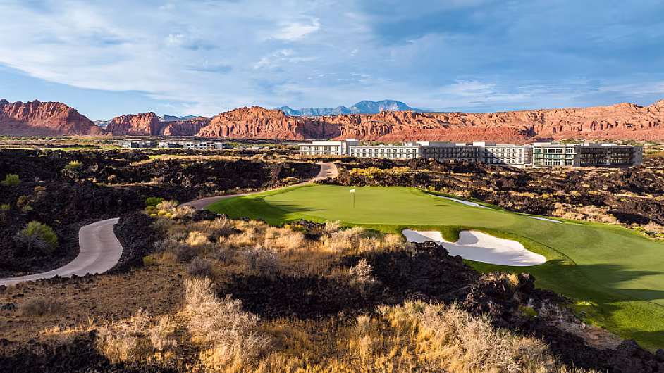A scenic golf course set in a desert landscape with red rock formations and mountains in the background, under a partly cloudy sky.