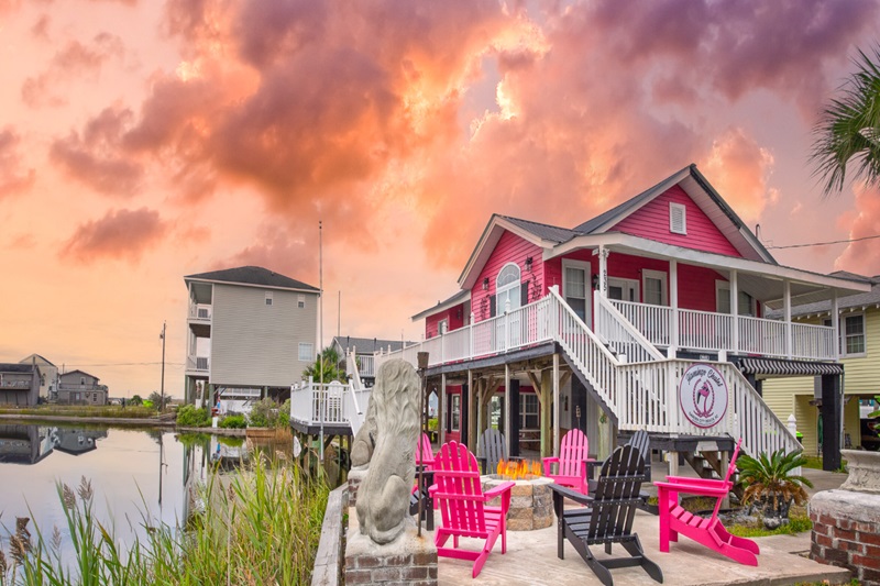 Flamingo Chalet Colorful pink and yellow beach house with a white deck, water views, and outdoor seating area at sunset.
