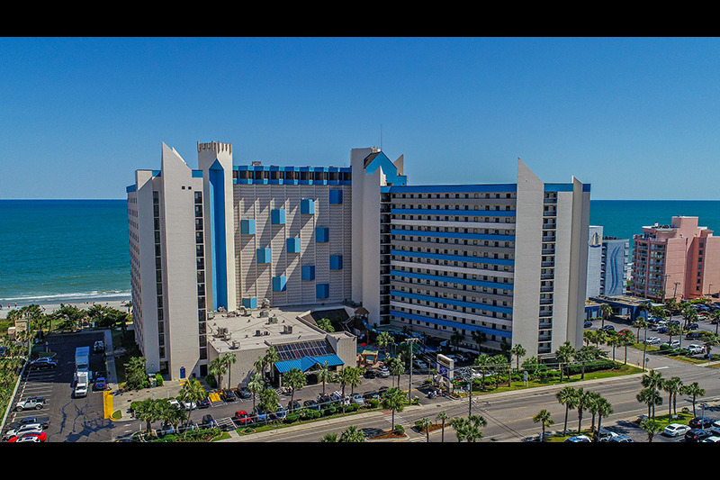 Ocean Reef A large beachfront hotel with modern architecture, surrounded by palm trees, parking lot, and ocean in the background at Myrtle Beach.