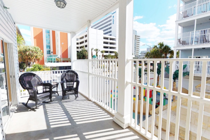 Myrtle Beach Villas - 105A Balcony of Myrtle Beach Villas with black wicker chairs, overlooking nearby buildings and palm trees near the beach.