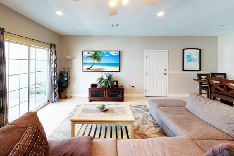 Myrtle Beach Villas - 303B Bright living room in Myrtle Beach Villas 303B with a large sectional sofa, TV, sliding glass doors, and tropical decor.