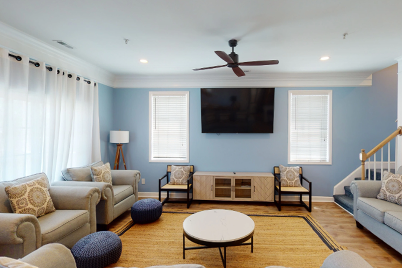 Bermuda Breeze - D Bright living room with blue walls, large windows with white curtains, beige sofas, a wall-mounted TV, and wooden flooring.