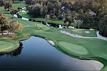 Aerial view of a golf course with green fairways, sand bunkers, water hazards, and trees, surrounded by residential houses.