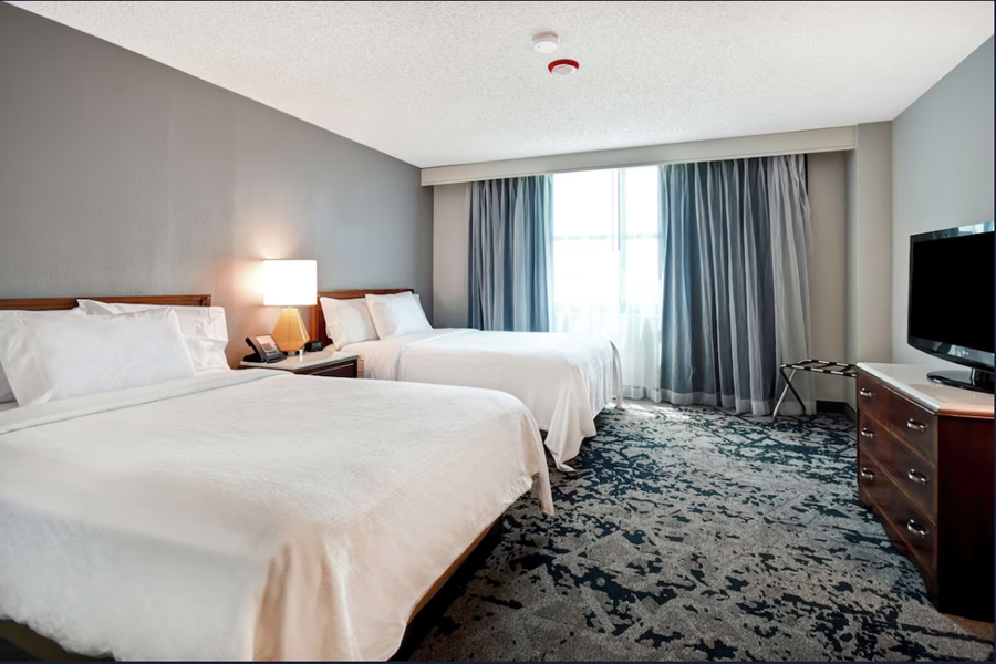 Interior of a hotel room with two double beds, a TV, a dresser, a lamp, and large windows with curtains, in Montgomery at Hilton Embassy Suites.