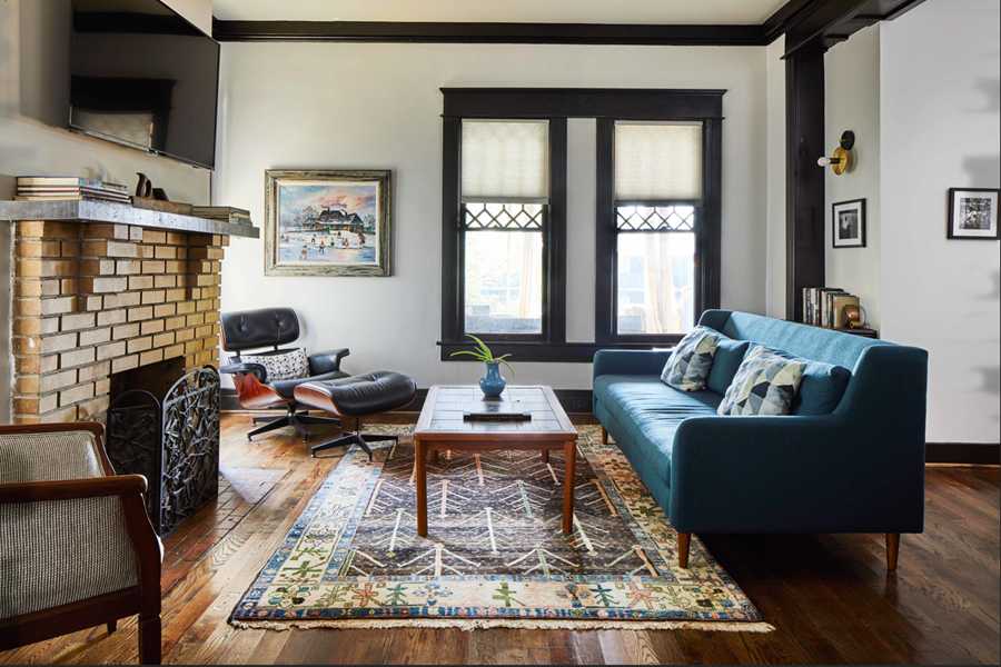 Interior of a stylish living room with a brick fireplace, blue sofa, black lounge chair, wooden coffee table, and large windows.