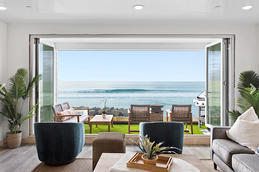 Bright living room with large open glass doors leading to a beachfront patio overlooking the ocean, with seating and a grill.