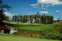 Scenic view of Otter Creek Golf Club with lush green fairways, sand bunkers, a small bridge, and a backdrop of trees under a blue sky.