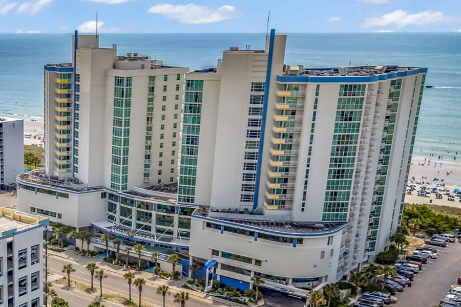 Avista Resort A tall, modern beachfront resort with glass balconies, overlooking the ocean and nearby sandy beach with umbrellas and people.