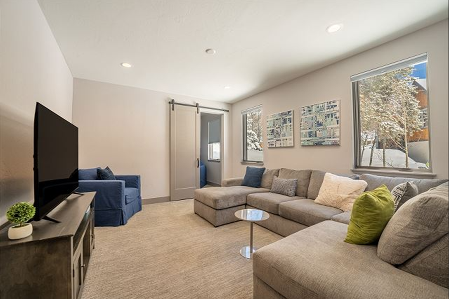 Bright living room with large windows showing snowy mountain scenery, featuring a beige sectional sofa and a blue armchair.