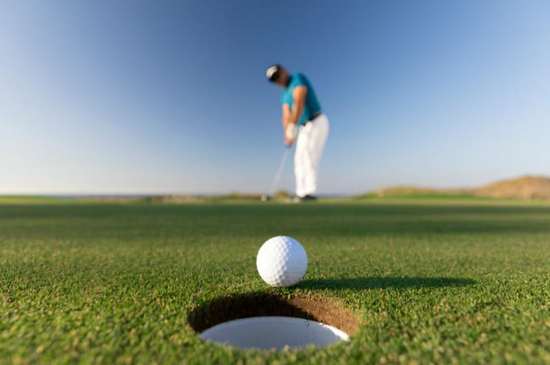 Close-up of a golf ball near the hole on a green, with a golfer preparing to putt in the background under a clear blue sky.