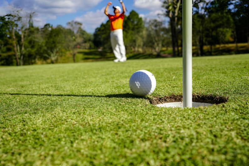 Close-up of a golf ball near the hole on a green, with a golfer celebrating in the background under a blue sky.