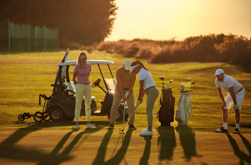 Golfers practicing on a lush green course during sunset, with golf carts and bags nearby, preparing for a game.