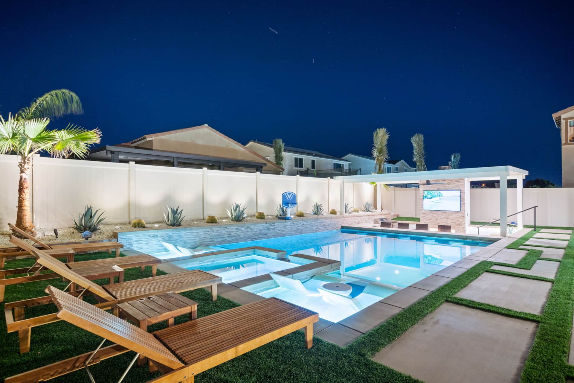 Coachella Luxe Oasis 6BR Resort Luxury resort backyard at night with illuminated pool, lounge chairs, palm trees, and outdoor TV, perfect for relaxing and entertainment.