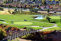 Scenic view of a golf course with players, a pond, and a bridge, surrounded by residential houses and colorful trees in the background.