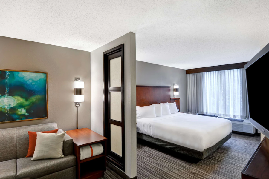 Modern hotel room at Birmingham Hyatt Place featuring a large bed, a sofa, artwork, and a window with sheer curtains.