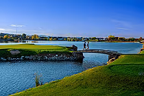 Scenic view of Pelican Lakes Resort with a golf course, a small bridge over water, and two people walking on the bridge.