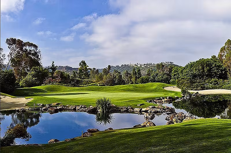Scenic golf course in San Diego with lush green fairways, water hazards, trees, and a backdrop of hills and blue sky.