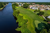 Aerial view of a golf course with lush green fairways beside a winding river, surrounded by residential houses and trees.