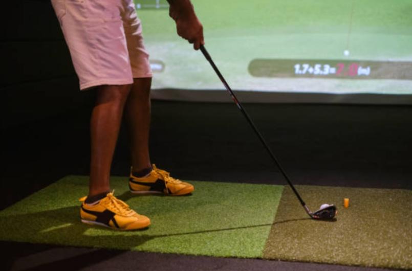 Person preparing to hit a golf ball indoors on a simulated golf course with a golf simulator, wearing yellow shoes and shorts.