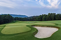 Scenic view of a golf course with a green, sand bunkers, trees, and distant hills under a partly cloudy sky.