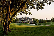 A scenic view of a golf course with a large house in the background, surrounded by trees and an American flag on a flagpole.