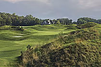 Scenic view of a golf course with lush green fairways, sand bunkers, and a house in the background, under a partly cloudy sky.