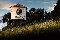 Water tower with Legends Golf & Country Club logo, surrounded by greenery and wildflowers, under a partly cloudy sky.