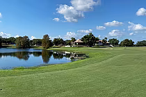 Scenic view of a golf course with a pond, lush green fairways, trees, and a clubhouse under a partly cloudy sky at MetroWest Golf Club in Orlando.