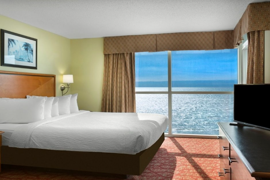 Bay View on the Boardwalk Hotel room with a large bed, ocean view through windows, and a flat-screen TV, offering a relaxing seaside vacation experience.