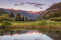 Scenic view of Vail Golf Club with players on the green, surrounded by lush trees, mountains, and a reflective pond at sunset.