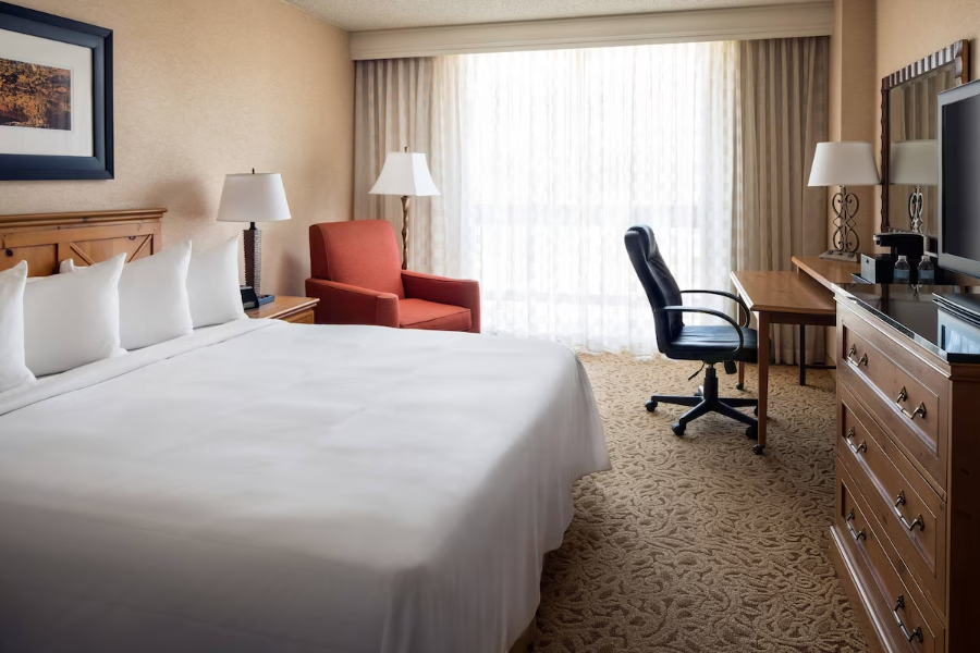 Elegant hotel room with king bed, armchair, desk, and TV, featuring neutral decor and large window with sheer curtains.