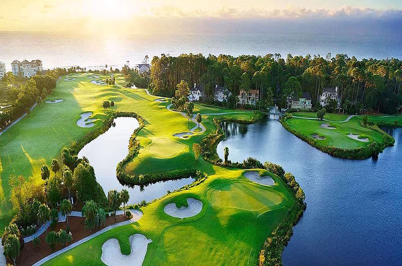 Aerial view of a lush golf course with water hazards, surrounded by trees and residential homes, overlooking the ocean at sunset.