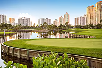 Scenic view of The Soffer golf course with lush green landscaping, a tranquil lake, and tall city buildings in the background.