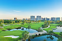Scenic view of a lush golf course with ponds, palm trees, and pathways, set against a backdrop of modern high-rise buildings.