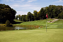 Scenic golf course with lush greens, a red flag on a putting green, and a tranquil pond bordered by trees under a blue sky.