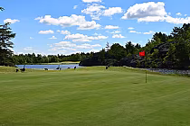 Scenic view of Pine Grove Golf Club with lush green, a red flag, serene lake, and trees under a blue sky in Sudbury.