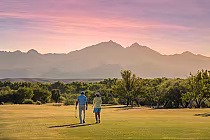 Couple walking on a lush golf course at Tubac Golf Resort, Arizona, with scenic mountain views and a colorful sunset sky.