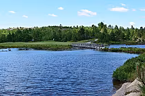 View of Stone Hill Golf Club with a serene lake, wooden bridge, and lush green trees under a clear blue sky.